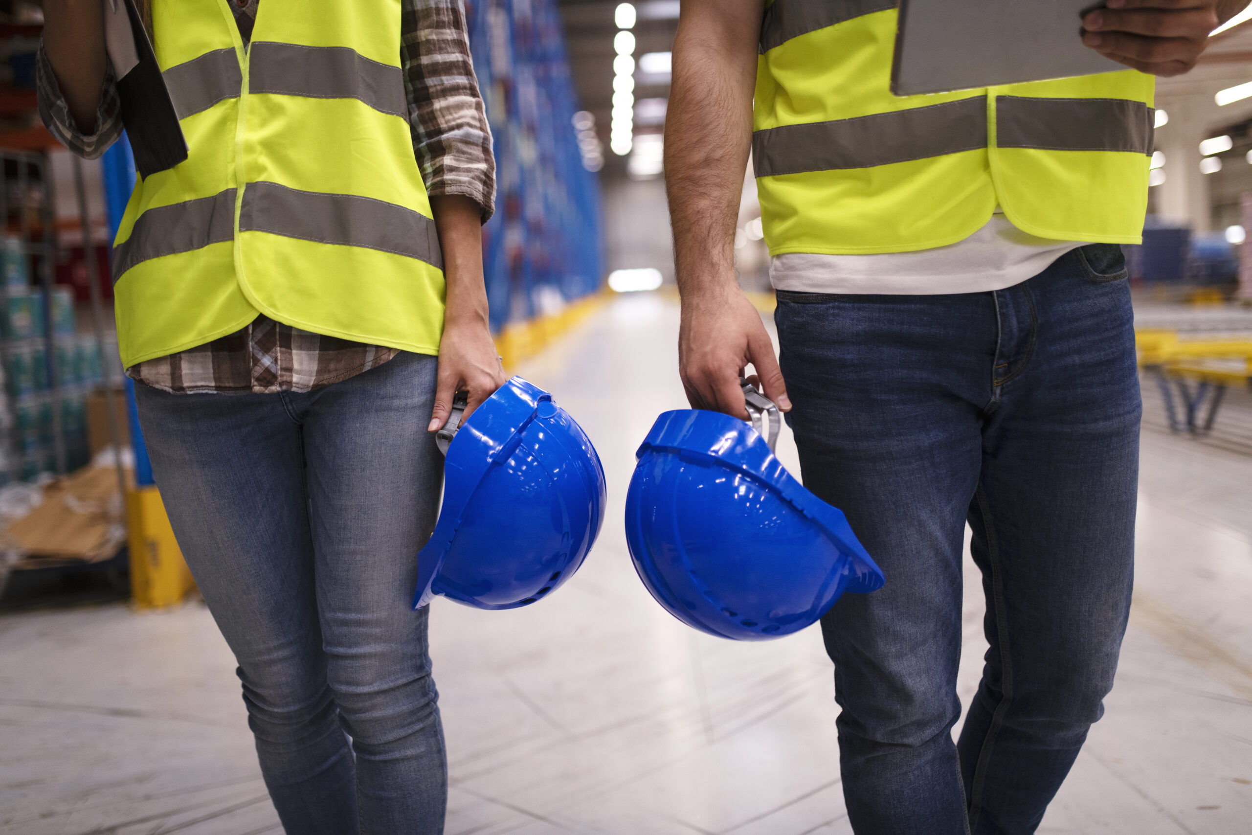 two unrecognizable workers reflective suit walking through warehouse holding blue protective hardhats scaled